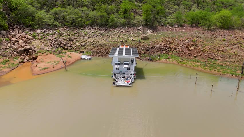 Drone shot of a large houseboat docked in the shallows of a river with a rocky river bank and vegetation in the background
