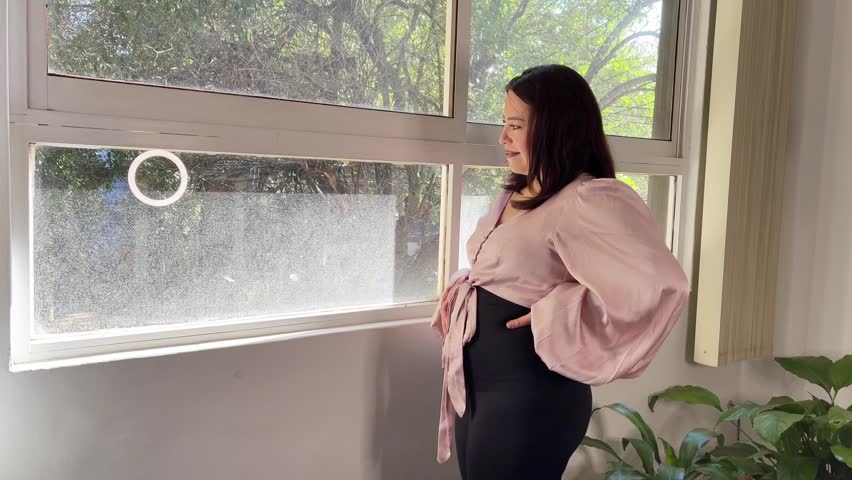 Hispanic pregnant entrepeneur holding belly and looking out window