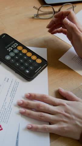 Vertical closeup of hands of unrecognizable young woman calculating domestic expenses using smartphone app, while examining overdue utility invoices