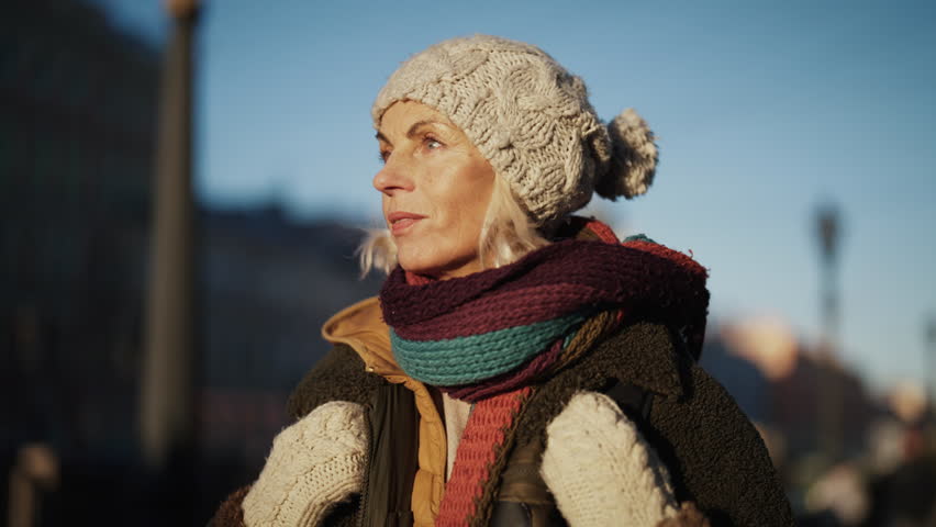 Aged woman tourist with backpack strolling in beautiful city, portrait outdoors. Inspired middle-aged lady walking in sunny weather, breathing frozen air, elderly female passer-by on street in winter