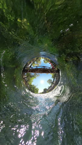 Vertical video. The camera transitions from a Tiny Planet effect to a wide-angle view, capturing the river flowing under a wooden bridge, surrounded by trees and greenery, with sunlight reflecting off