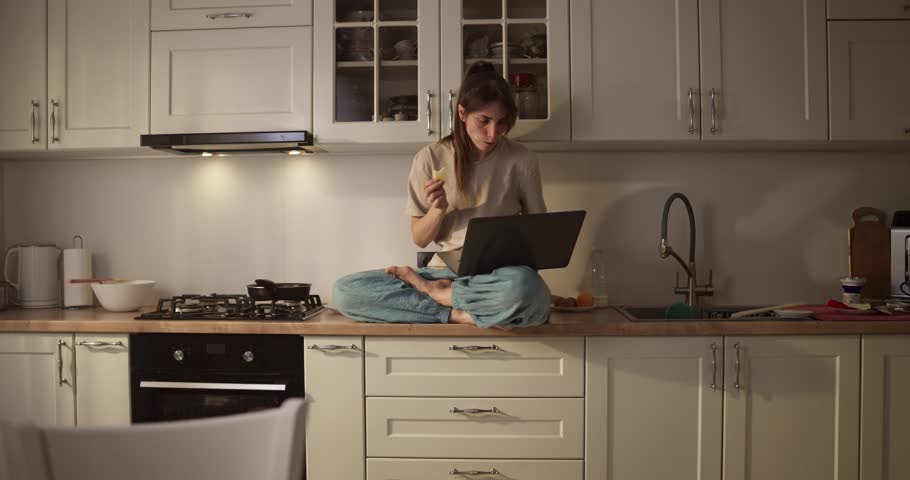 Focused girl sitting at the table eating breakfast and working using a laptop in a modern kitchen in the morning