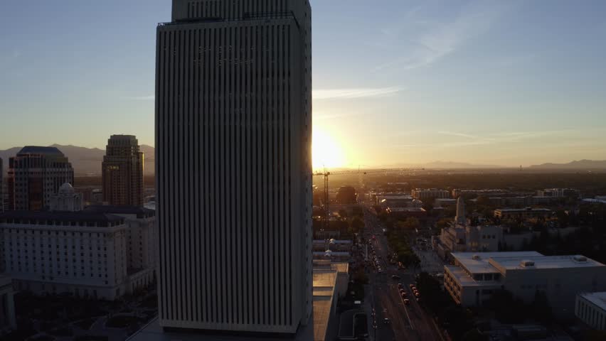 Aerial drone shot of the west Salt Lake Valley from downtown, trucking right over skyscrapers, homes, and busy roads. Vibrant fall trees glow in the golden-orange hues of a colorful autumn sunset.