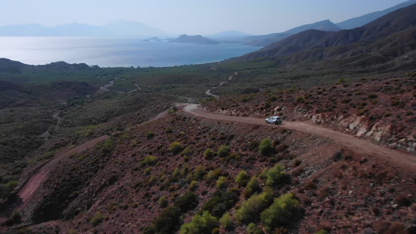 Winding mountain road with car driving along dusty and unpaved road, overlooking Karaincir Bay, surrounded by rugged terrain, Turkey. Aerial forward