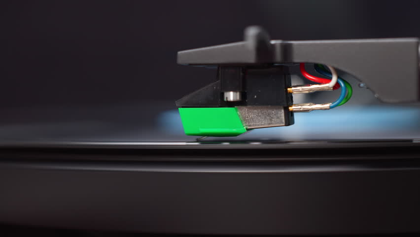 A macro shot of a record player needle touching down on a black vinyl record's lead-in groove. Crisp details capture the stylus and tonearm in a static profile view.