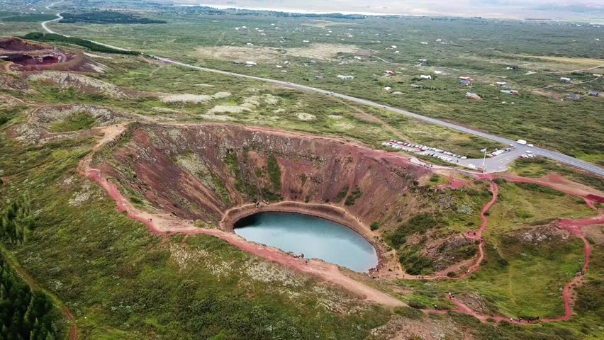 Kerid Volcanic crater lake reflecting blue sky in Iceland Golden Circle, featuring red slopes, green moss and hiking paths, aerial view from above, drone pulling out