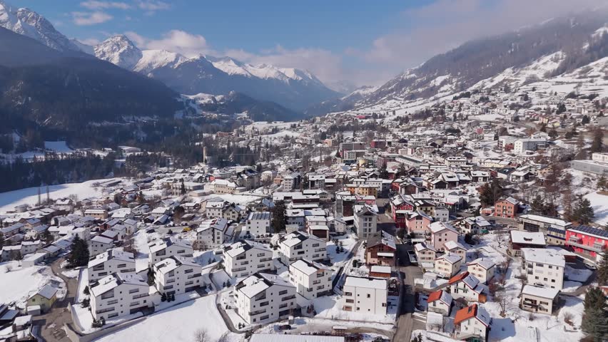 Picturesque swiss city during Sunny winter day. Modern Apartment houses and homes with alp panorama in distance. Aerial wide shot Snow-covered Buildings ion Scuol Town, Switzerland.