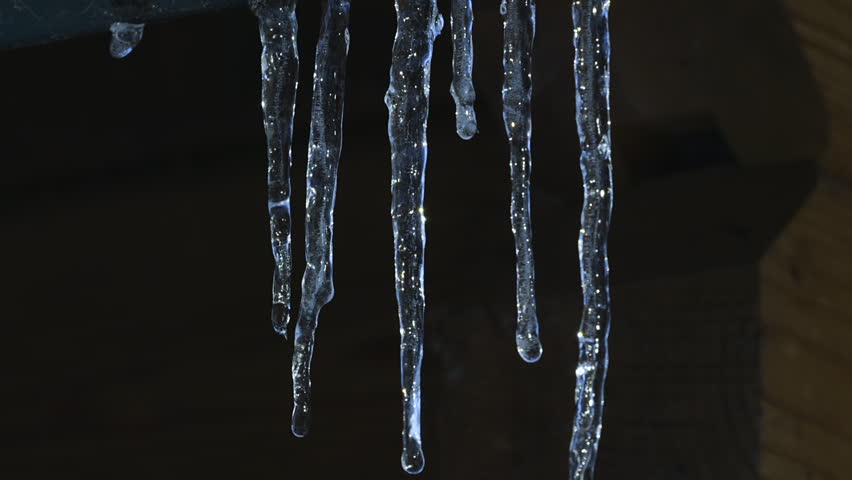 Panning top to bottom of a small row of icicles melting near a wooden cabin wall. Closeup.