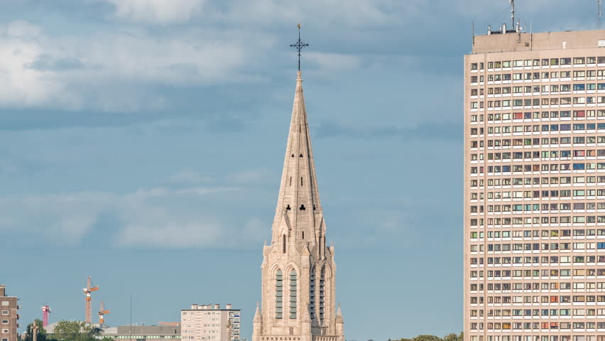 Aerial timelapse of Saint Servatius Church in Schaerbeek, Brussels, Belgium. Neo-Gothic spire surrounded by red-roofed houses under a blue cloudy sky. High-rise buildings and cranes in the background