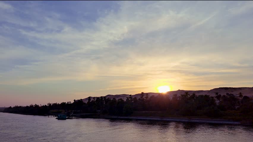 A serene sunset over the Nile River with a cruise ship in the distance. The sky is painted with hues of orange and purple, reflecting on the calm waters. Aswan to Luxor, Egypt.