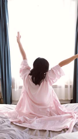 Happy Asian woman on bed awakened from enough and healthy sleep. Woman stretching in bed after wake up, back view.