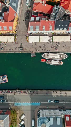 Vertical top down drone shot tracking along the iconic canal of Nyhavn, Copenhagen, Denmark. Showcasing its colourful roofs, historic boats and many pedestrians walking along the waterfront.