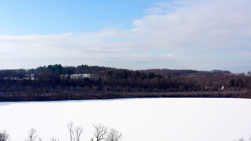 Aerial drone view flying in over the frozen Mohansic Lake in FDR Park, Yorktown Heights, NY. The vast snowy expanse contrasts with the winter forest and distant rolling hills.