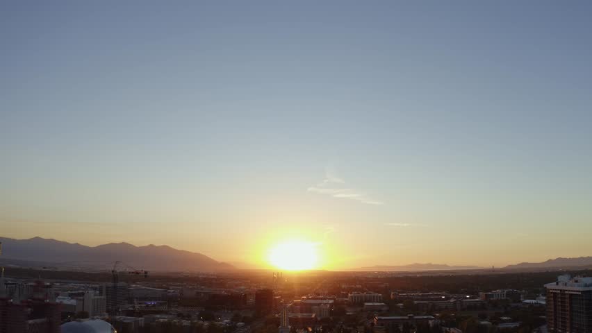 Aerial drone shot of the west Salt Lake Valley from downtown, trucking left over skyscrapers, homes, and busy roads. Vibrant fall trees glow in the golden-orange hues of a colorful autumn sunset.