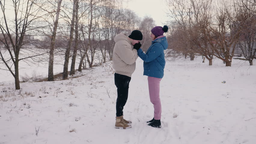 Young couple standing face to face in light falling snow, sharing a tender moment in a scenic winter landscape surrounded by bare trees. High quality 4k footage