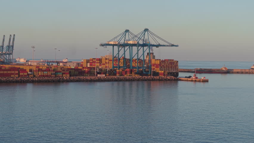 Aerial view of a busy container port with large shipping cranes and stacked cargo containers. The golden light of sunset illuminates the maritime trade hub in Las Palmas, Gran Canaria.