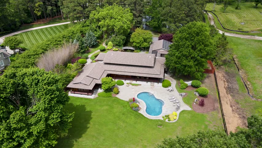 A cinematic drone slowly backs away from a stunning modern house, revealing a vibrant backyard with lush green grass, a bright blue pool, colorful flower bushes, and a dock extending over the water