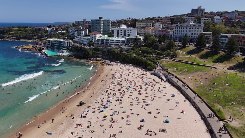 Drone shot of Bondi Beach, showcasing people swimming, sunbathing, and relaxing along the coastline. Panning aerial view, NSW, Australia