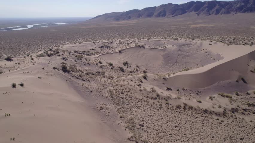 A large sand dune in the middle of the desert. Top view from a drone. A mountain of sand on which various dry bushes grow. A strong wind is blowing, lifting the sand. Blue sky and white clouds