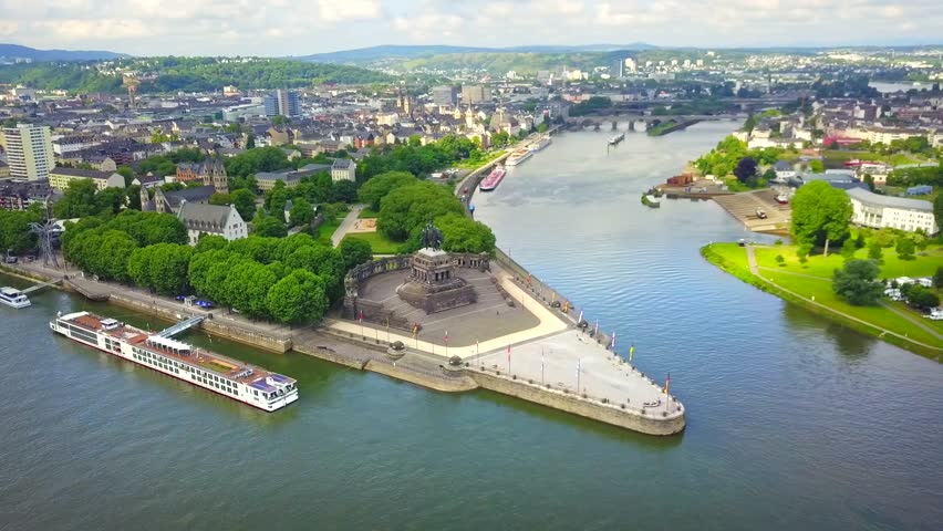Aerial view of Deutsches Eck in Koblenz, Germany, where the Rhine and Moselle rivers merge. The historic monument, lush greenery, and passing cargo boats create a dynamic and scenic riverside scene.