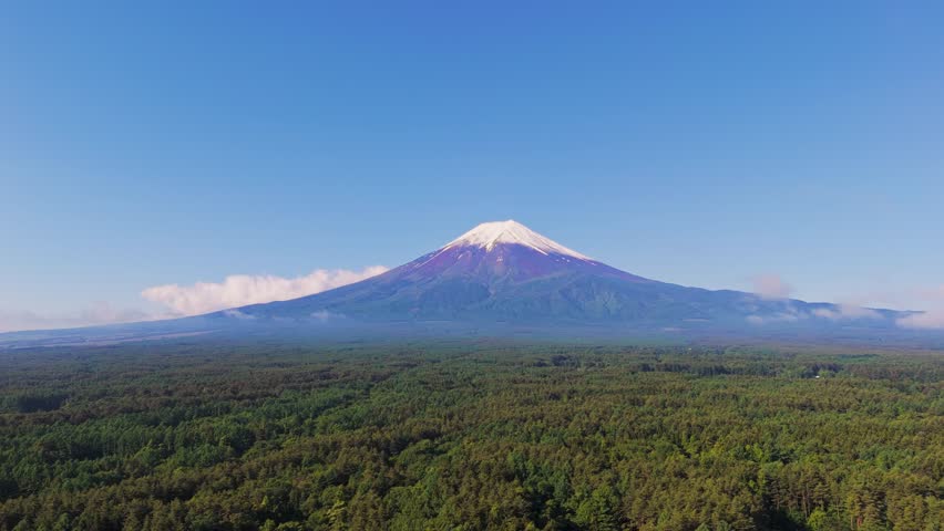 Mount Fuji rises above Kawaguchiko town in a peaceful morning scene. Wispy clouds float across the mountain slopes, complementing the lush green forest and bright blue sky