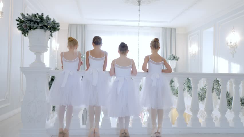 Four young girls in white ballet dresses are standing on a balcony. The girls are wearing bows and are looking out over a balcony. Scene is one of elegance and grace