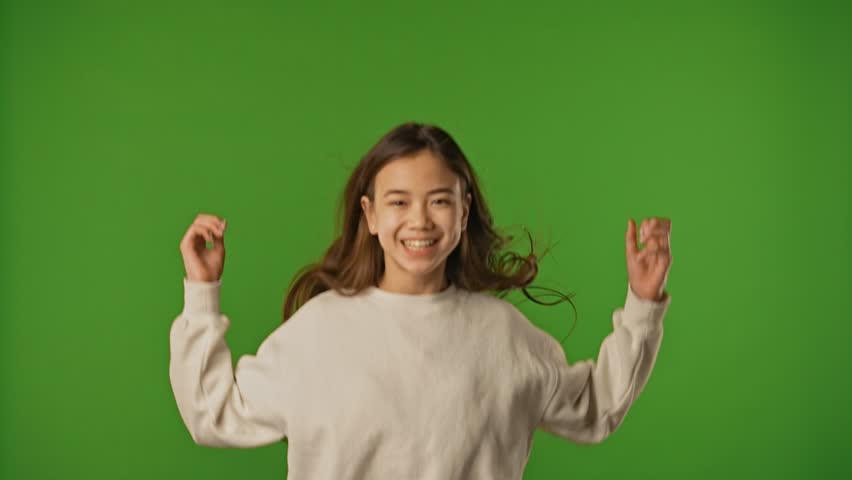Asian girl child in shirt jumping up in scene and smiling looking at camera, positive expression, isolated on chroma key green screen studio background. Slow motion.