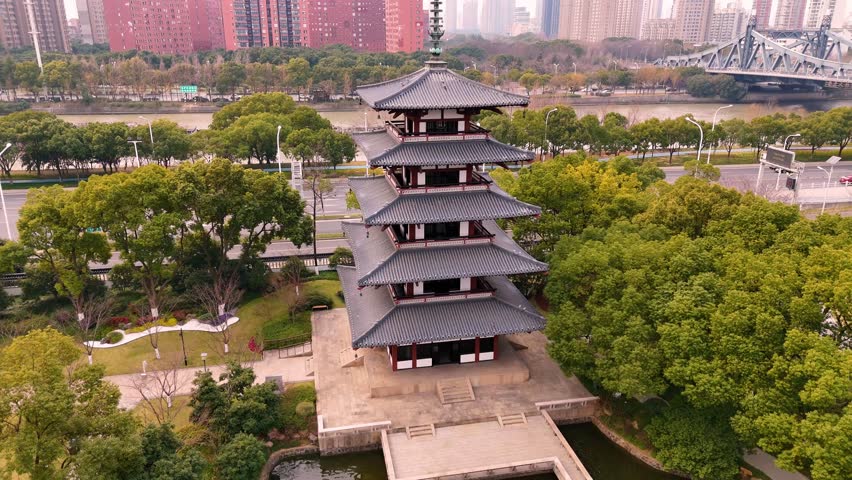 Aerial drone footage of a serene urban park with a small lake and a traditional Chinese pagoda. The video captures lush greenery, tranquil water reflections, and the harmonious blend of nature and arc
