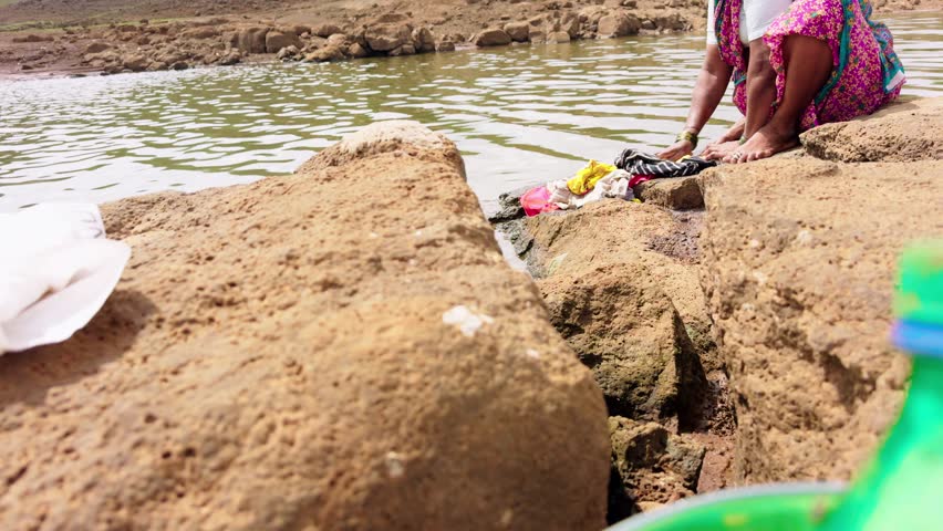 Villagers wash cloth against hard stones near river, Rural ladies washing clothes on rocky ground beside lake, Woman cleans garments over coarse rock by stream, People soak laundry upon jagged surface