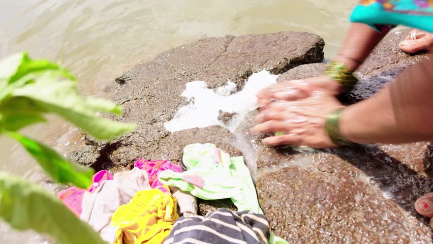 Rural ladies washing clothes on rocky ground beside lake, Villagers wash cloth against hard stones near river, Woman cleans garments over coarse rock by stream, People soak laundry upon jagged surface