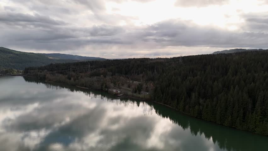 Tranquil lake reflects a cloudy sky, bordered by a dense evergreen forest, creating a peaceful and scenic landscape. British Columbia, Canada.