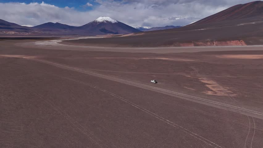 Aerial view of a white pickup truck kicks up dust as it moves along a rugged trail in the remote Atacama desert. Snowy peaks and arid, volcanic terrain stretch into the distance