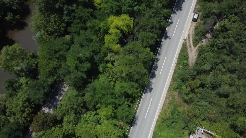 Aerial shot of a black car speeding down a road through the forest. On the left is a river, on the right is a parked gray car. Tracking shot
