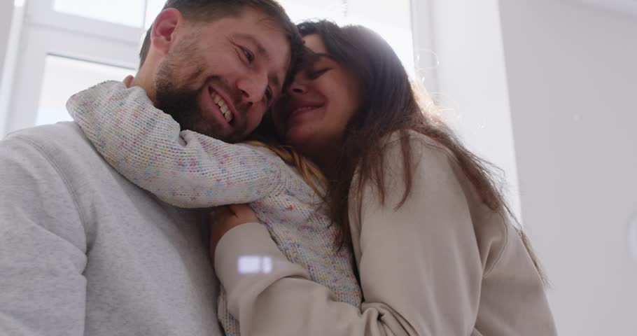 Cheerful smiling happy mother and father hugging their cute little child girl standing in living room at home by the window in sunlight. Happy family concept. 4k video. Slow motion video.