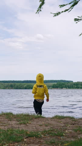 Kid wearing a yellow sweatshirt and black pants stands at the lake. Baby boy throws the pebbles into the water. Vertical video.