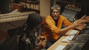 Mature sound engineer explaining audio production details to African American musician, both sitting at desk with mixing console in professional recording studio. High angle view - Powered by Shutterstock - Get 15% off with code: PIKWIZARD15