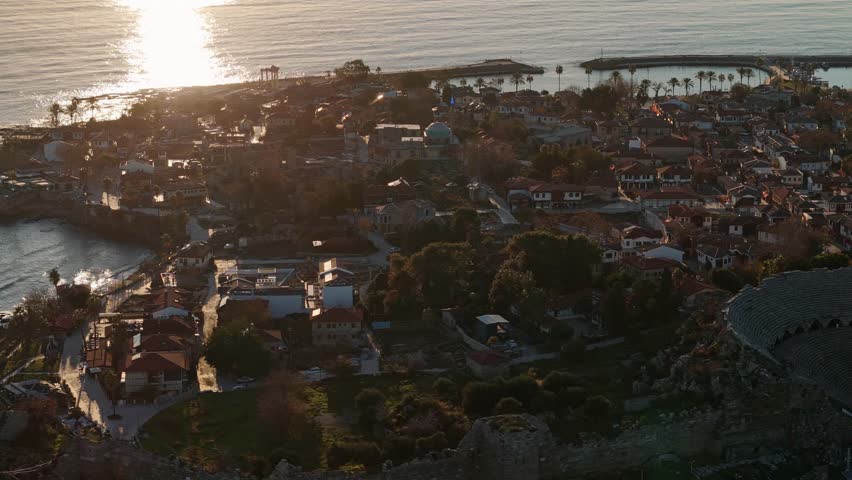 Aerial perspective of a Mediterranean coastal town. The ruins of an amphitheater dominate the scene, with traditional houses and a harbor visible along the shoreline. Side Ancient City