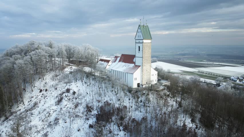 Drone footage of a snowy winter landscape as it moves away from a hilltop church, revealing a serene snow-covered forest in the foreground. A peaceful and cinematic winter scene.