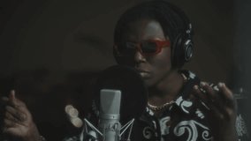 Young African American male singer in headphones, patterned shirt and trendy sunglasses singing expressively into microphone as performing in music recording studio - Powered by Shutterstock - Get 15% off with code: PIKWIZARD15