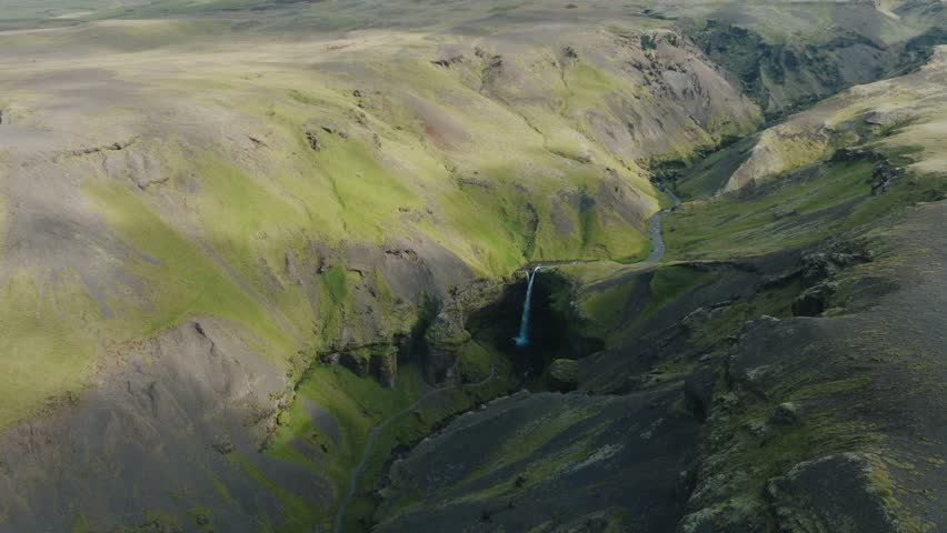Wide aerial shot of Kvernufoss waterfall in a vast green valley in Iceland. The scene captures the dramatic cliffs, the winding trail, and the serene beauty of untouched Nordic nature.