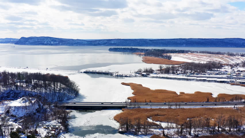 Scenic drone fly-in over Croton Bay in Croton-On-Hudson, NY, showcasing the snow-covered landscape, winding expressway, train tracks, and viaduct under a partly cloudy winter sky.