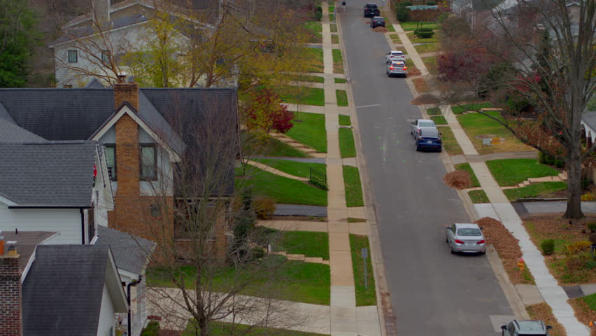 High-quality aerial shot over a picturesque street in Ladue, St. Louis, Missouri, during autumn. Captures vibrant fall foliage and a car pulling out and driving away. Perfect for suburban themes.