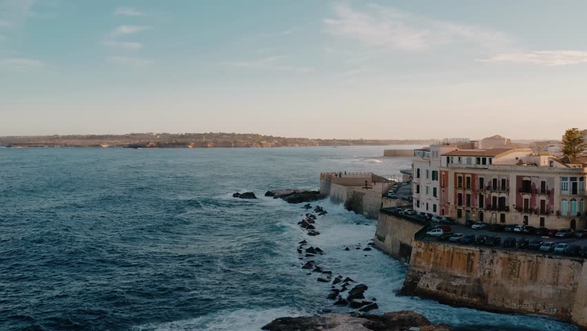 Aerial drone shot of Ortigia island along the Mediterranean sea cost with large waves crashing on the fortress. Historic charming medieval town in Sicily at sunset.