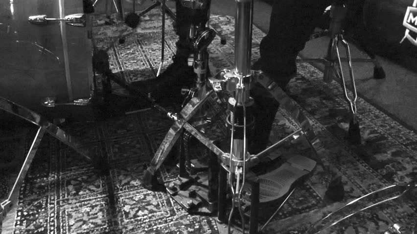 Grayscale footage of a persons legs playing acoustic bass drum with double bass pedal in slow motion in a studio room where a stylish carpet in on the ground. Hi hat and other drum equipment visible.