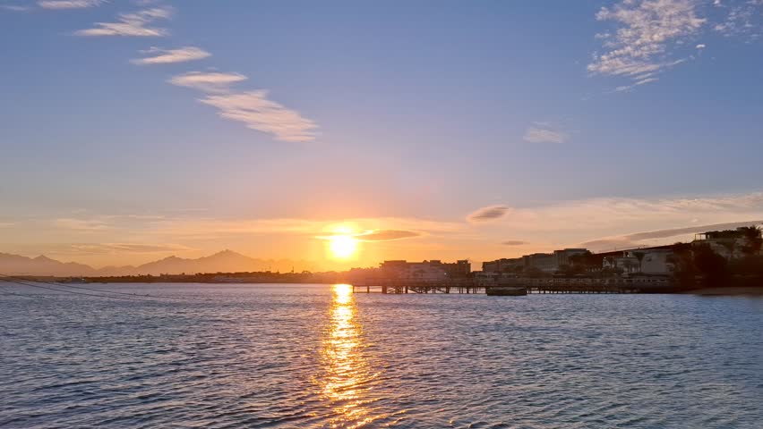 A stunning Red Sea sunset with orange and blue skies, reflecting on calm blue waters, in Hurghada, Egypt. Luxury yachts and distant buildings enhance the scenic resort view.