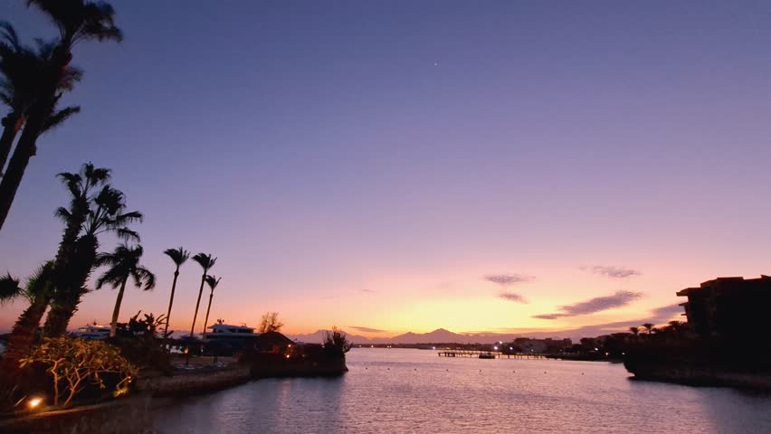 A stunning Red Sea sunset with orange and blue skies, reflecting on calm waters, in Hurghada, Egypt. Silhouetted hotels and yachts at the port create a serene resort scene.