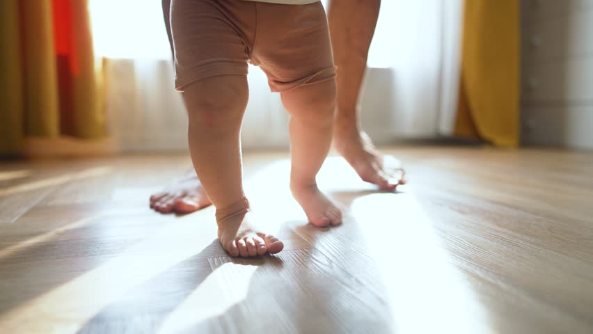 Father helps take first step. Son learning to walk with father guidance. Proud father supports son first walk indoors. Son takes first step while walking with. Father teaches son to step and walk.