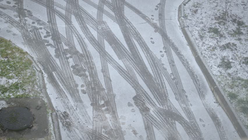 A winter scene featuring a snow-covered road with multiple intersecting tire tracks and footprints. The contrast between the white snow and dark imprints creates an abstract pattern