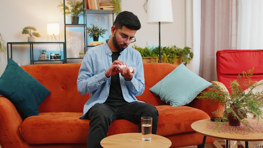 Indian young man taking medicine pill with water sitting on cozy sofa at home. Hispanic ill sick guy takes medication prescribed by his physician. Medicine, health care, pharmacy and people concept.
