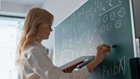 Teacher writing math equations on chalkboard looking book closeup. Woman educator solving mathematical tasks on blackboard classroom. Blonde female tutor in glasses teaching algebra in high school. - Powered by Shutterstock - Get 15% off with code: PIKWIZARD15